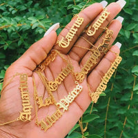 A hand holds several gold necklaces with gothic script zodiac signs like Aquarius, Scorpio, and Libra against a background of green leaves.