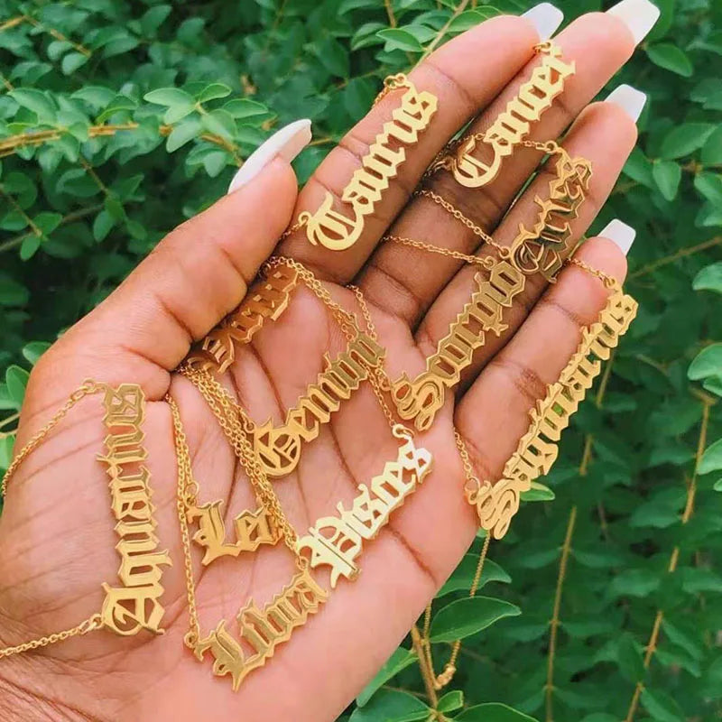 A hand holds several gold necklaces with gothic script zodiac signs like Aquarius, Scorpio, and Libra against a background of green leaves.