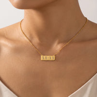 A close-up of a model's neck and upper chest wearing a delicate gold chain necklace with a small rectangular gold pendant engraved with angle number 11:11.