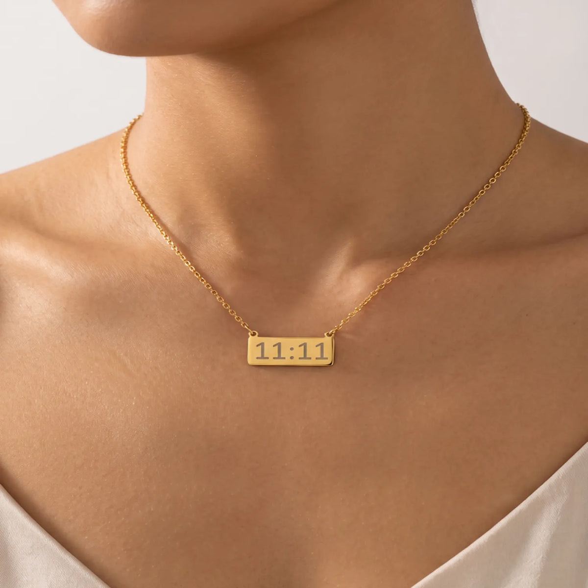 A close-up of a model's neck and upper chest wearing a delicate gold chain necklace with a small rectangular gold pendant engraved with angle number 11:11.