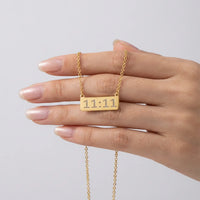 A gold bar necklace engraved with angel number 11:11 held on a person's fingers against a light gray background.
