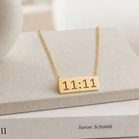 A gold bar necklace with angel number 11:11 engraved on a gold chain, displayed on a light gray fabric book stand with a book underneath.