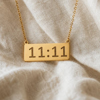 A gold rectangular pendant necklace engraved with angel number 11:11 in a warm, polished finish, hanging from a delicate gold chain against a textured beige linen fabric background.