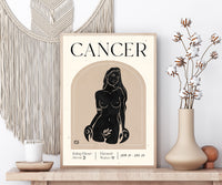 A framed art print featuring a black ink illustration of a Cancer zodiac sign on a cream background, styled on a wooden shelf next to a macrame wall hanging, a candle, and a vase with dried cotton stems.