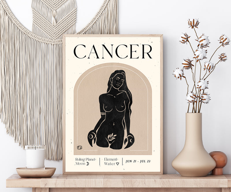 A framed art print featuring a black ink illustration of a Cancer zodiac sign on a cream background, styled on a wooden shelf next to a macrame wall hanging, a candle, and a vase with dried cotton stems.
