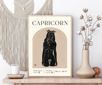 A framed Capricorn art print displayed on a wooden shelf in a styled room setting, flanked by a macrame wall hanging, a candle, and a vase with dried cotton stems.