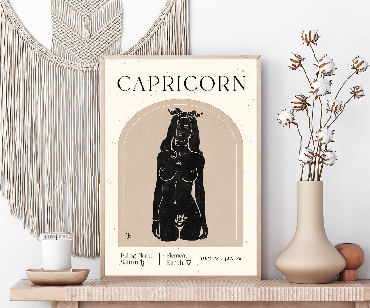 A framed Capricorn art print displayed on a wooden shelf in a styled room setting, flanked by a macrame wall hanging, a candle, and a vase with dried cotton stems.