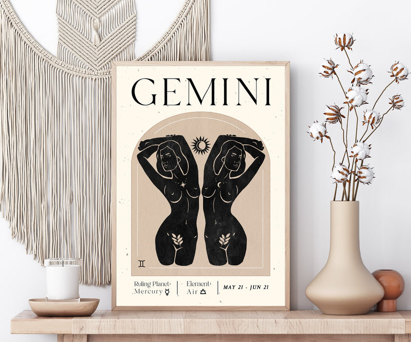 A framed Gemini astrology art print displayed on a wooden shelf in a styled room setting, flanked by a macrame wall hanging, a candle, and a vase with dried cotton stems.
