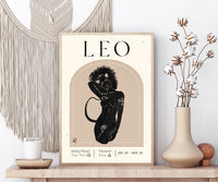A framed art print featuring a stylized black illustration of a Leo zodiac sign on a beige background, styled on a wooden shelf next to a macrame wall hanging, a candle, and a vase with dried flowers.