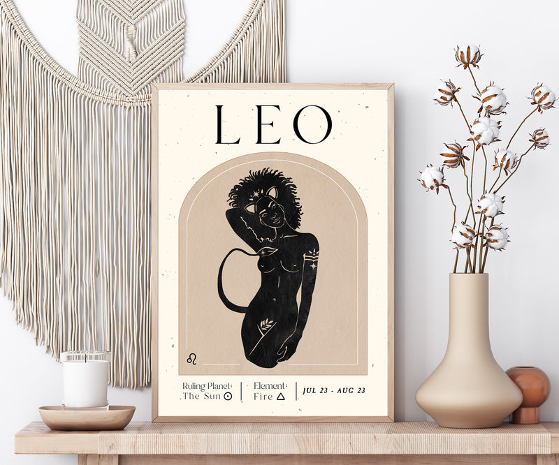 A framed art print featuring a stylized black illustration of a Leo zodiac sign on a beige background, styled on a wooden shelf next to a macrame wall hanging, a candle, and a vase with dried flowers.