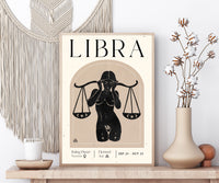 A framed Libra zodiac art print with a black silhouette illustration of a woman holding scales, styled on a wooden shelf with a macrame wall hanging, candle, and dried cotton stems in a vase.