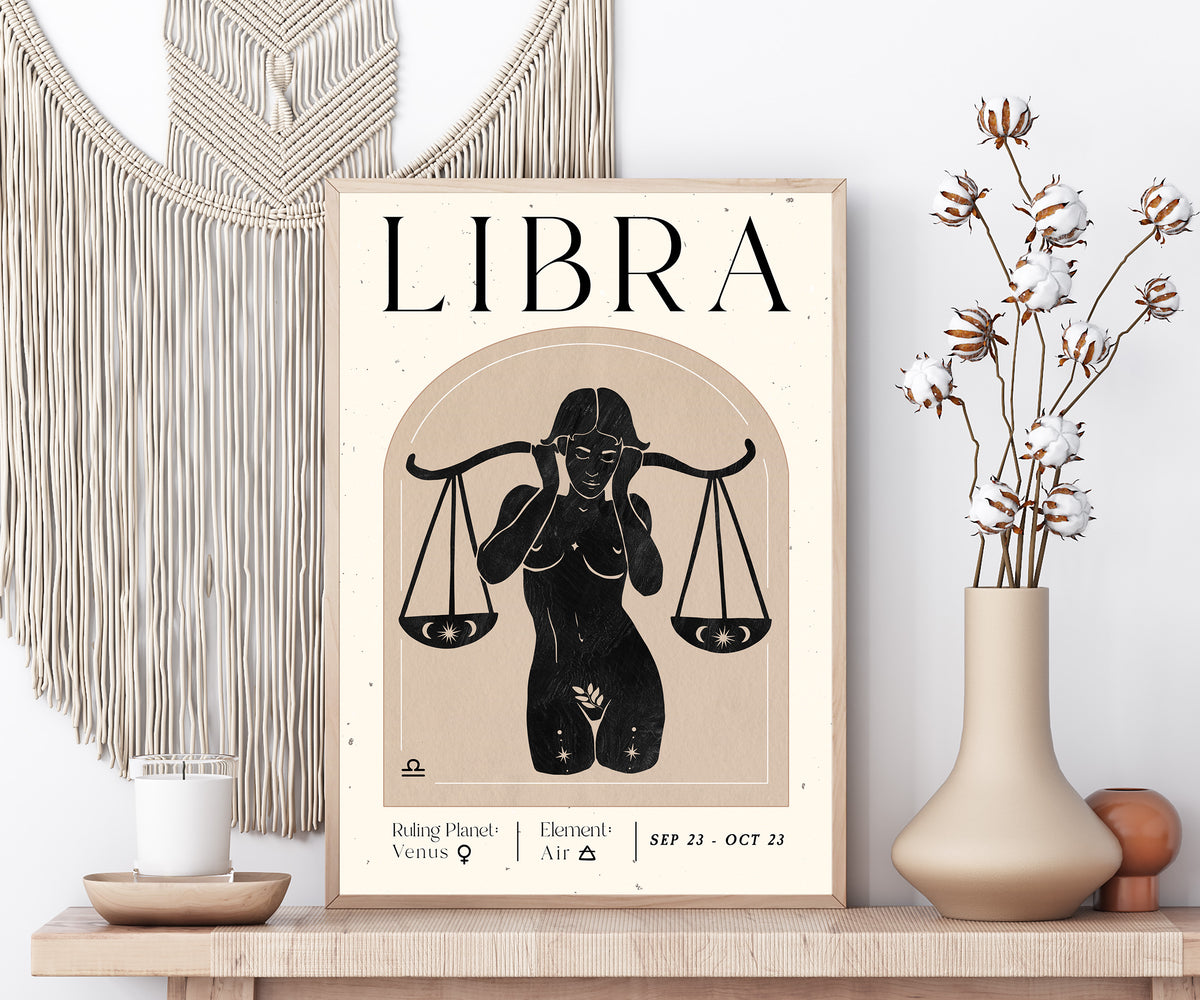 A framed Libra zodiac art print with a black silhouette illustration of a woman holding scales, styled on a wooden shelf with a macrame wall hanging, candle, and dried cotton stems in a vase.