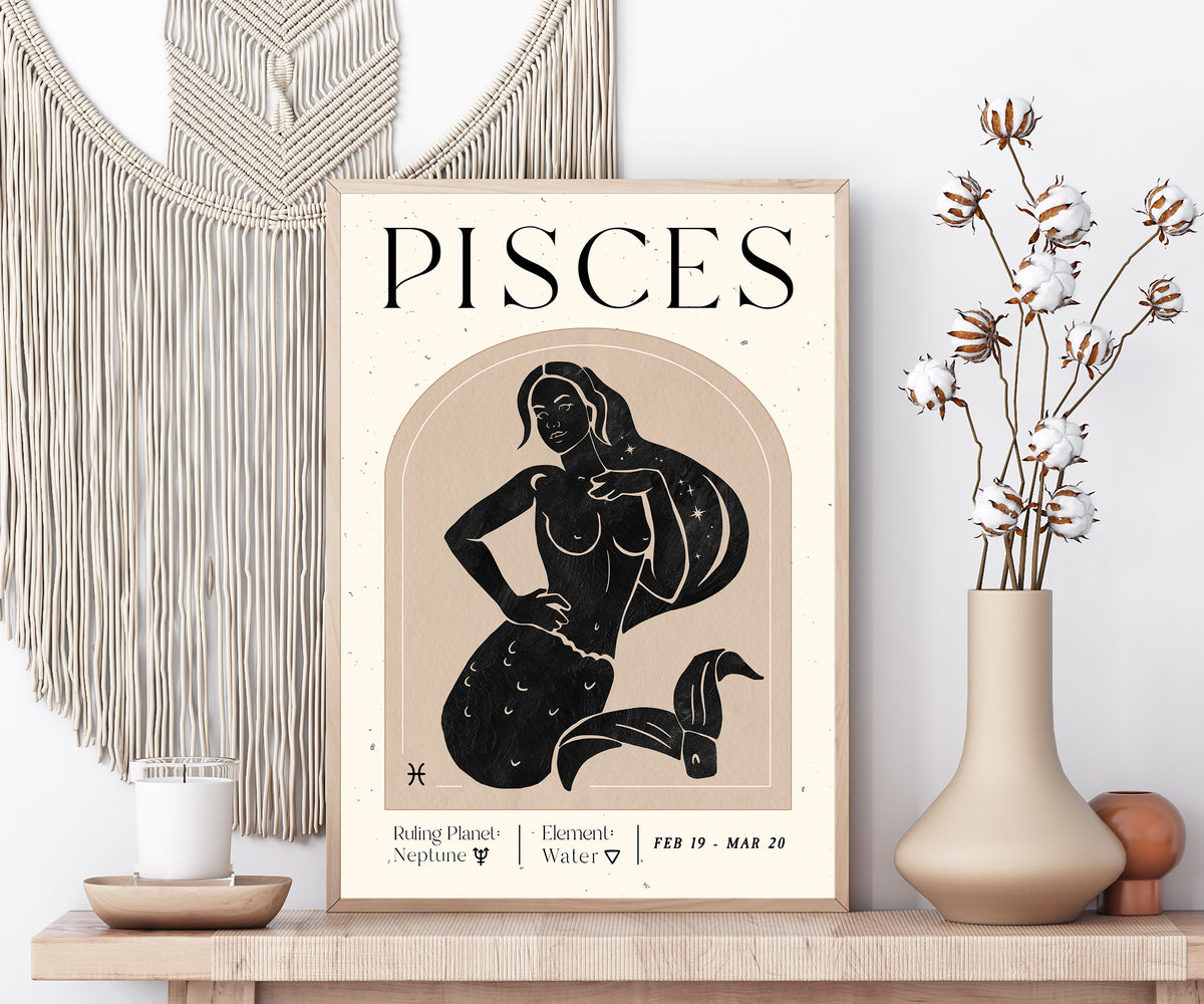 A framed Pisces zodiac art print displayed on a wooden shelf in a styled home decor setting, featuring a black line art illustration of a mermaid on a beige background with cream text.