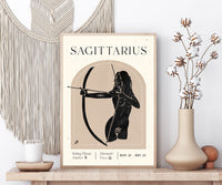 A framed Sagittarius astrology poster displayed on a wooden shelf, styled with a macrame wall hanging, a candle, and a vase of dried flowers.