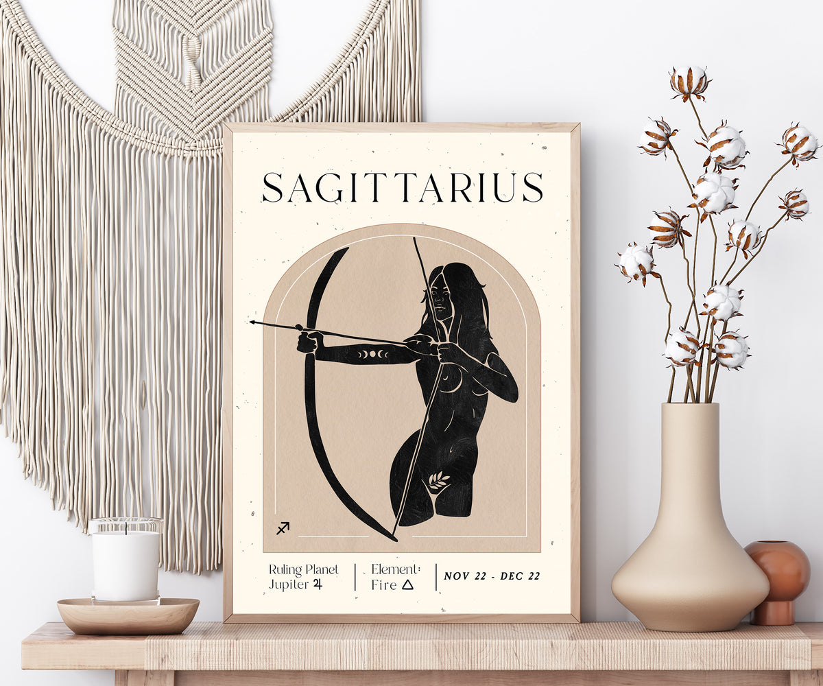 A framed Sagittarius astrology poster displayed on a wooden shelf, styled with a macrame wall hanging, a candle, and a vase of dried flowers.