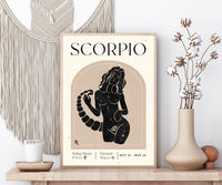 A framed art print featuring a black and beige Scorpio zodiac sign poster styled on a wooden shelf next to a macrame wall hanging and a vase with dried cotton stems.