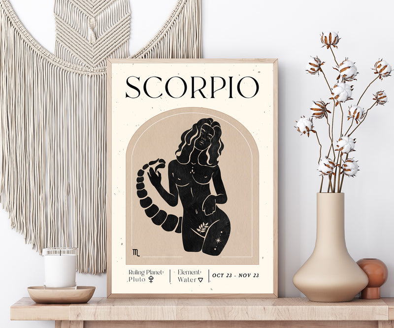 A framed art print featuring a black and beige Scorpio zodiac sign poster styled on a wooden shelf next to a macrame wall hanging and a vase with dried cotton stems.