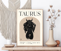 A framed Taurus astrology poster displayed on a wooden shelf, styled with a macrame wall hanging, a candle, and a vase of dried cotton stems.