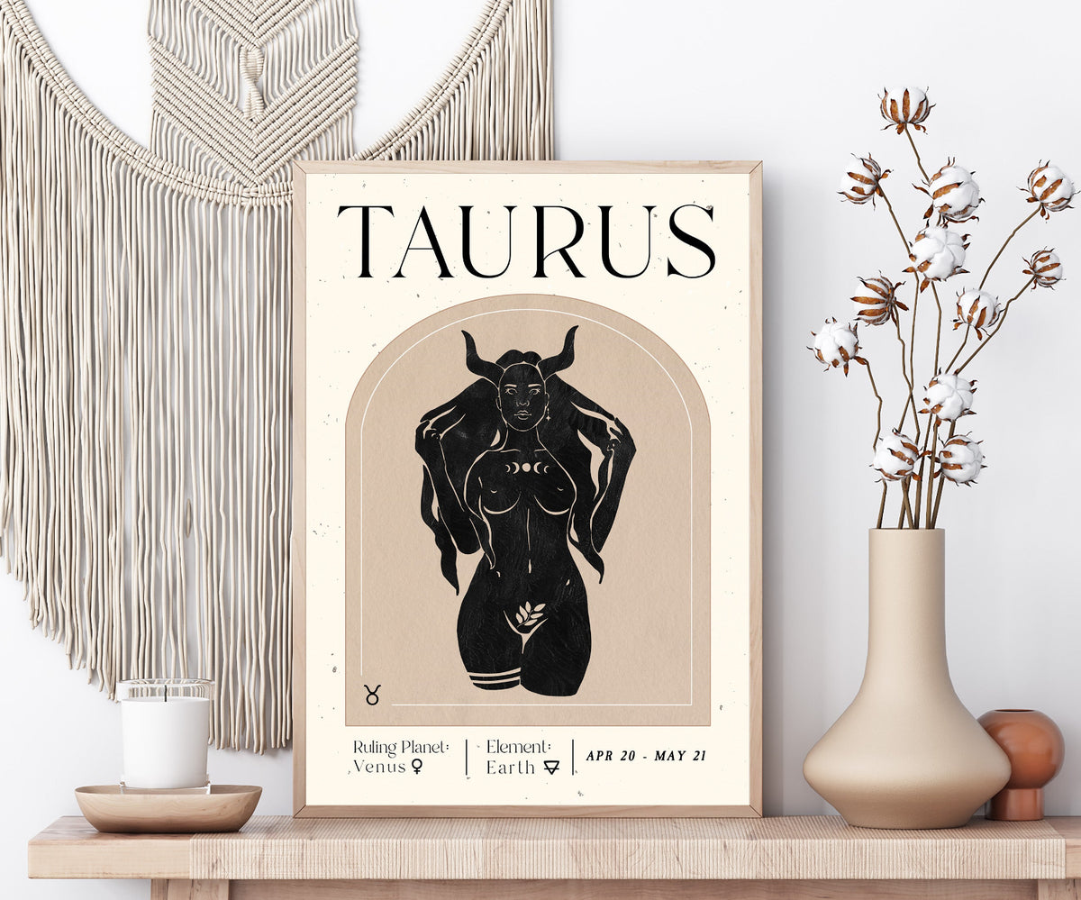 A framed Taurus astrology poster displayed on a wooden shelf, styled with a macrame wall hanging, a candle, and a vase of dried cotton stems.