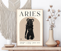 A framed Aries zodiac poster displayed on a wooden shelf, styled with a macrame wall hanging, a candle, and a vase with dried flowers.