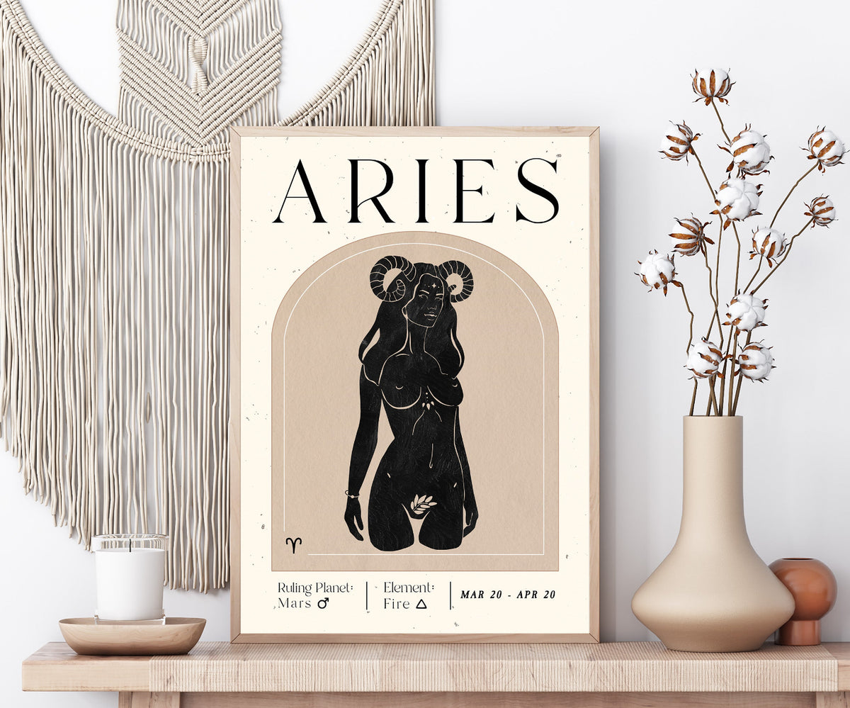 A framed Aries zodiac poster displayed on a wooden shelf, styled with a macrame wall hanging, a candle, and a vase with dried flowers.