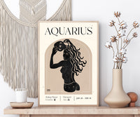 A framed art print featuring a retro Aquarius zodiac poster with a black silhouette of a woman and cream text on a beige background, styled on a wooden shelf with a macrame wall hanging, a candle, and a vase with dried flowers.