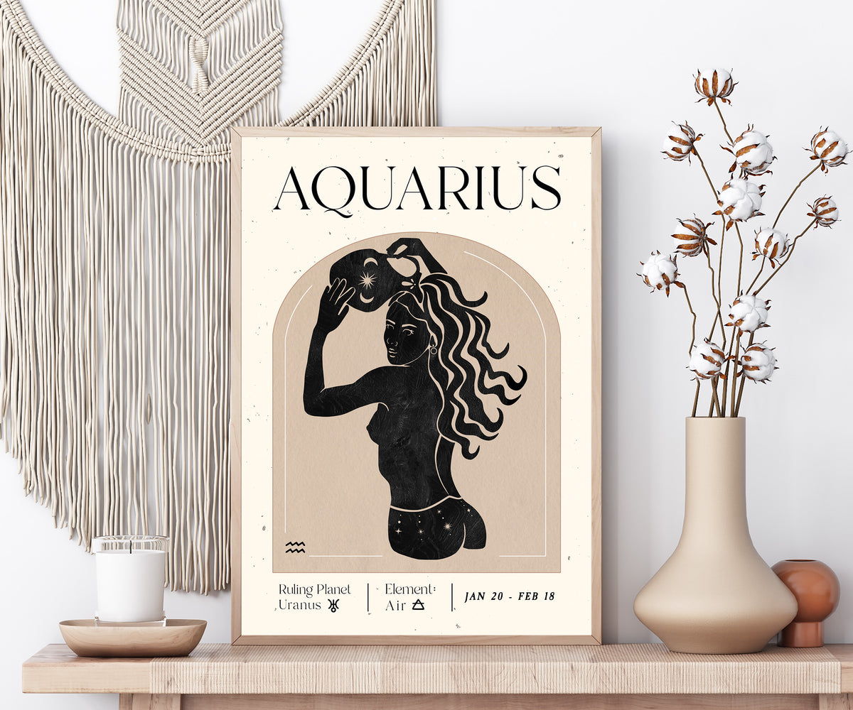 A framed art print featuring a retro Aquarius zodiac poster with a black silhouette of a woman and cream text on a beige background, styled on a wooden shelf with a macrame wall hanging, a candle, and a vase with dried flowers.