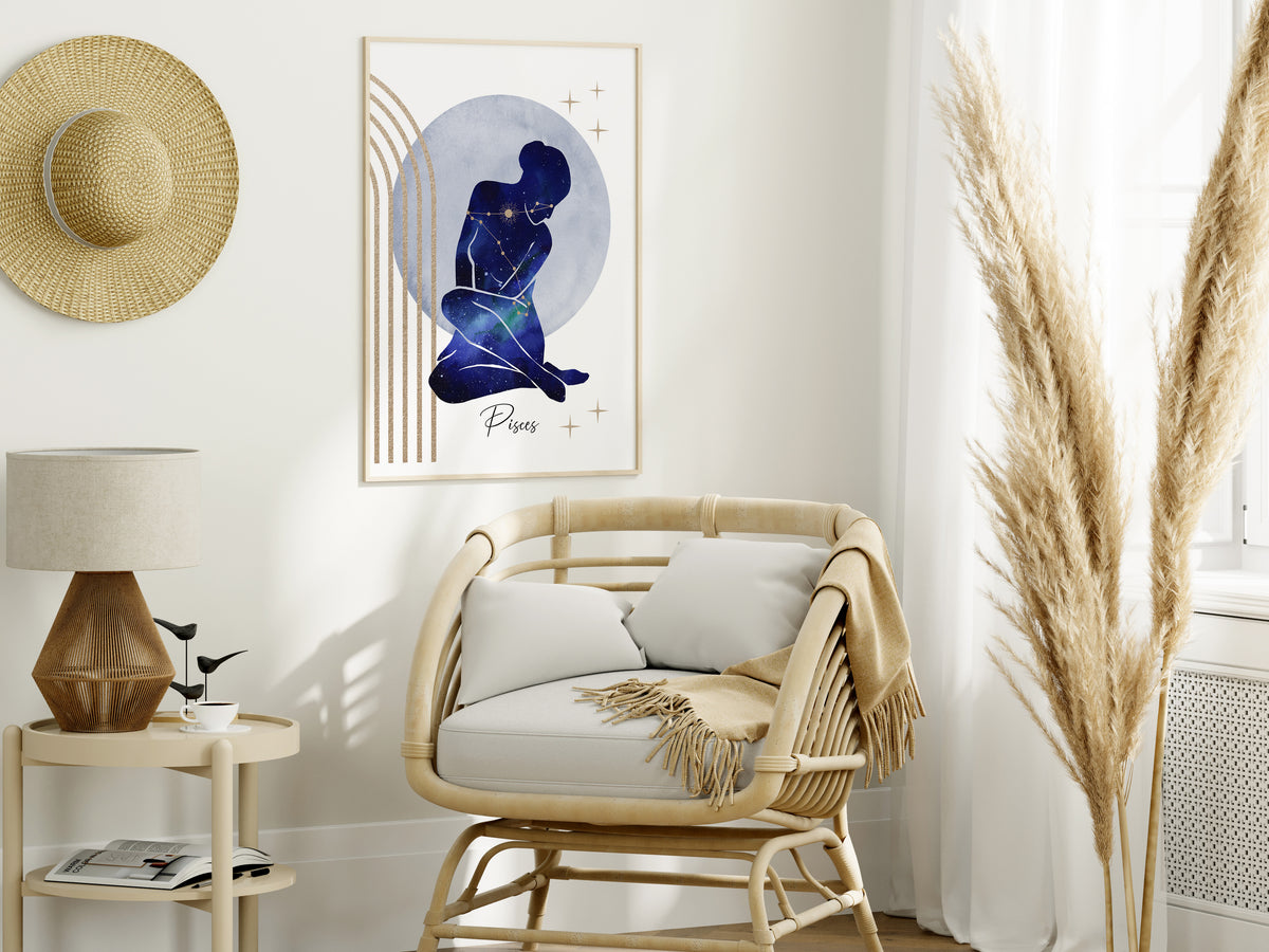 A framed art print featuring a dark blue silhouette of a seated figure against a pale blue circular background, styled in a bright, modern room next to a rattan armchair and a woven straw hat.