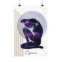A vertical art print featuring a stylized seated Capricorn figure filled with a purple and blue galaxy pattern, set against a pale blue circular backdrop with gold stars, arches, and the word 'Capricorn' at the bottom. The print is shown hanging from two black clips against a white background.