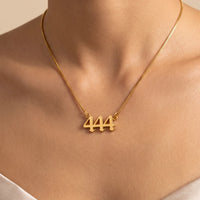 A close-up of a model's neck and upper chest wearing a delicate gold box chain necklace with a small gold pendant featuring the number 444.