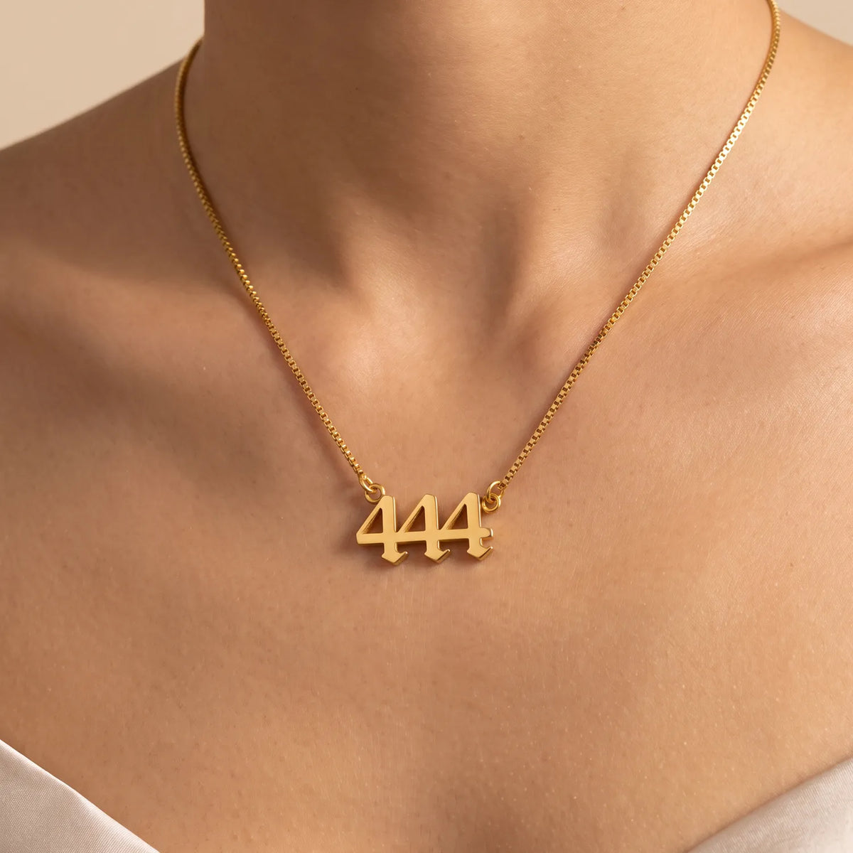 A close-up of a model's neck and upper chest wearing a delicate gold box chain necklace with a small gold pendant featuring the number 444.