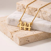 A gold box chain necklace with a 444 number pendant, displayed on stacked beige travertine and white marble blocks.