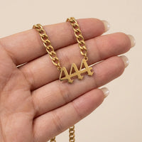 A gold curb chain necklace with a stylized '444' pendant held in a hand against a light beige background.