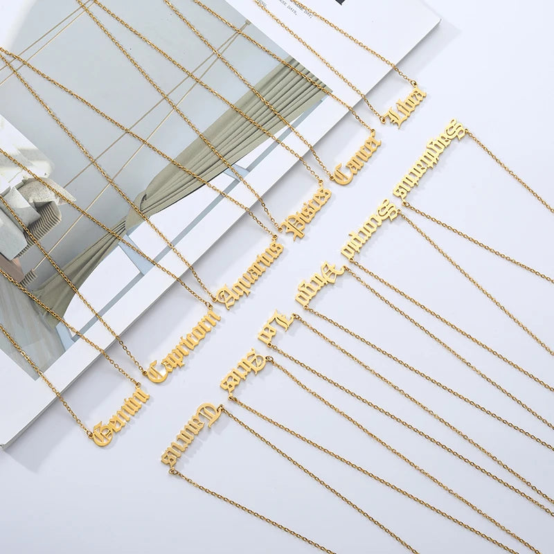A set of gold necklaces with gothic script zodiac signs laid out on a white surface with a magazine page underneath, 