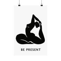 A minimalist black and white art print featuring a silhouette of a person in a yoga pose with the text 'BE PRESENT' below, shown hanging from two black clips against a white background.