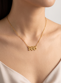 A close-up of a model wearing a delicate gold chain necklace with an angel number pendant representing 888 styled with a soft cream-colored top against a neutral light gray background.