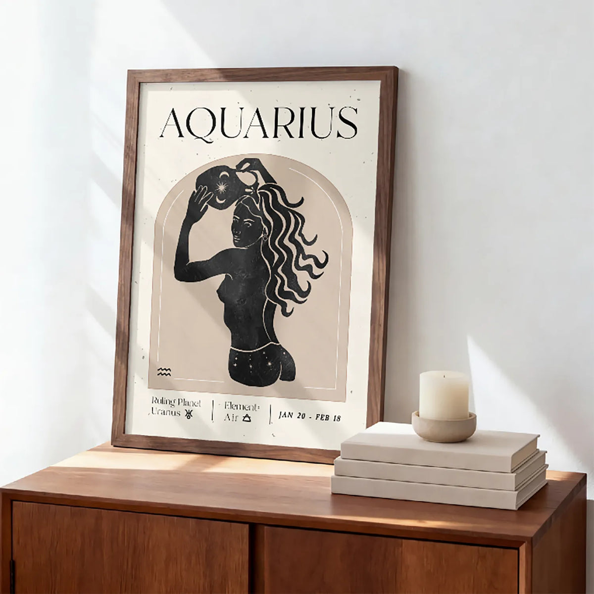 A framed Aquarius zodiac art print with a black silhouette illustration on a cream background, leaning against a white wall on a wooden cabinet next to stacked books and a candle.