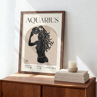 A framed Aquarius zodiac art print with a black silhouette illustration on a cream background, leaning against a white wall on a wooden cabinet next to stacked books and a candle.