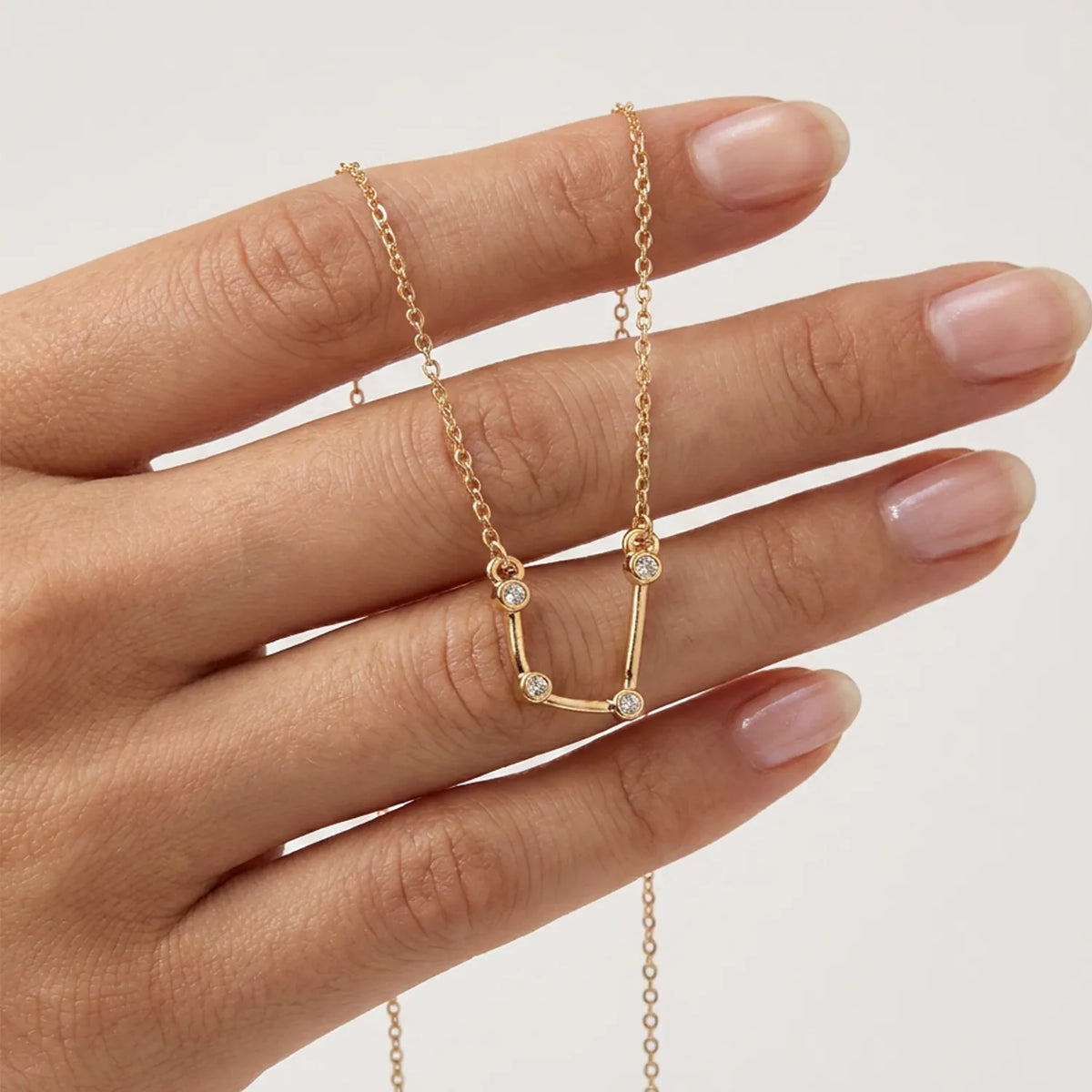A delicate gold necklace with an aquarius constellation sign shown on a person's hand against a light neutral background. 