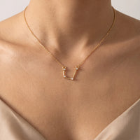 A delicate gold necklace with an aquarius constellation pendant set with tiny clear stones, shown worn on a model's neck and upper chest.