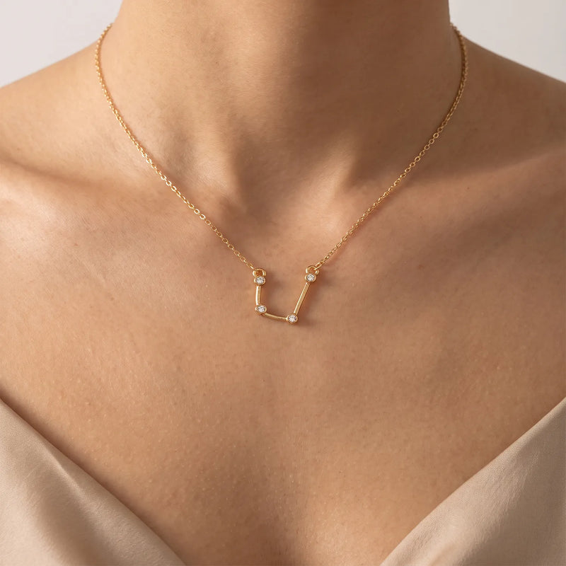 A delicate gold necklace with an aquarius constellation pendant set with tiny clear stones, shown worn on a model's neck and upper chest.