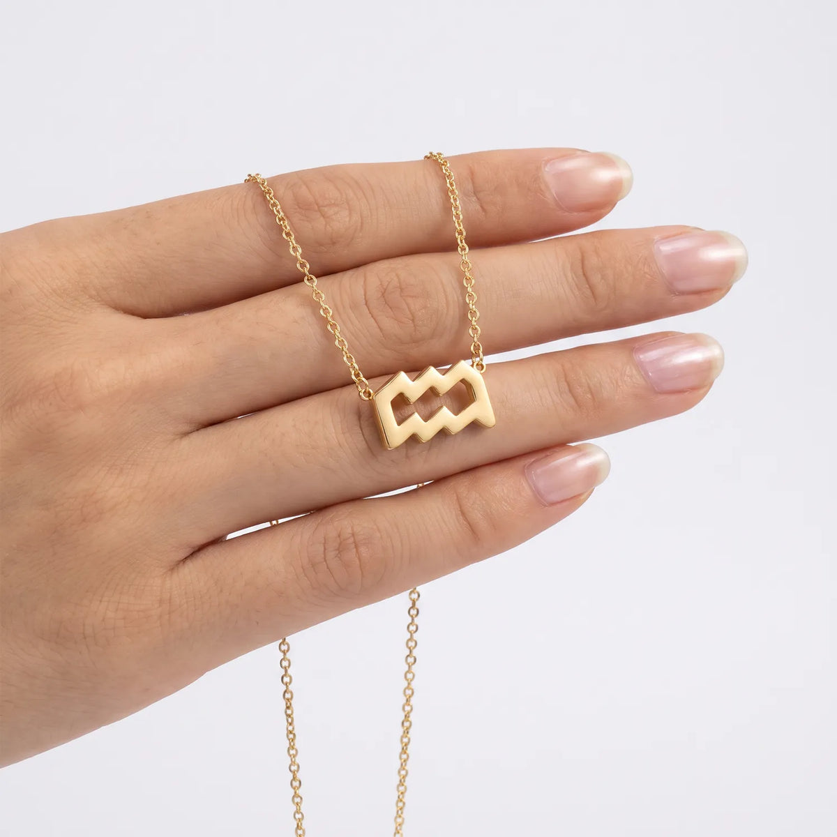 A gold necklace with an aquarius pendant is draped over a person's hand against a clean white background