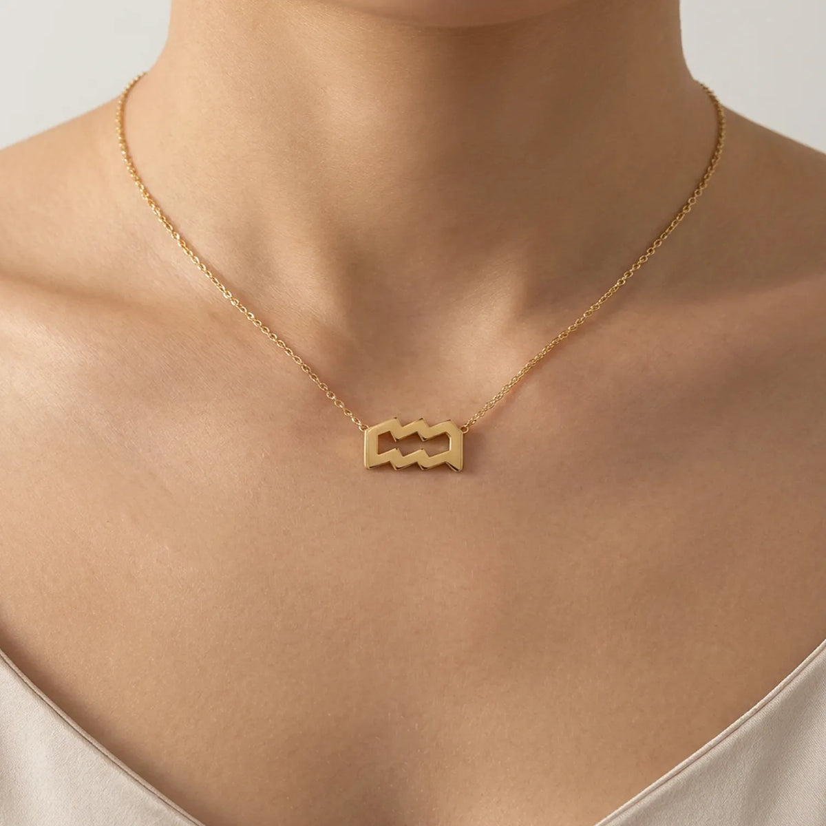 A close-up of a model's neck and upper chest wearing a delicate gold chain necklace with an aquarius sign pendant