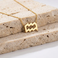 A gold necklace with an aquarius pendant resting on stacked beige stone tiles.