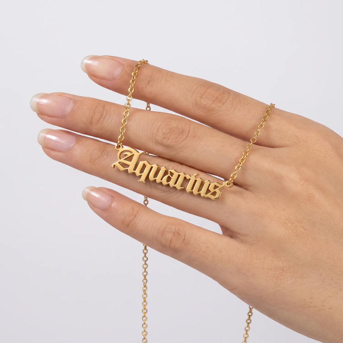 A gold nameplate necklace spelling 'Aquarius' in gothic lettering, shown worn on a person's ring finger against a light gray background.