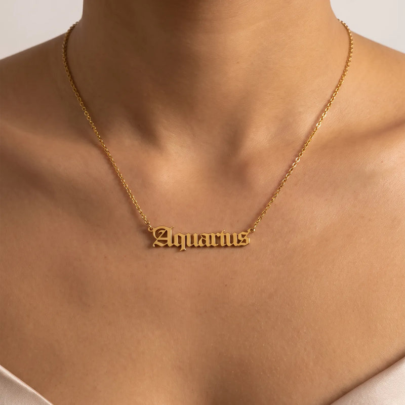 A gold nameplate necklace spelling 'Aquarius' in a gothic script worn on a model's neck, shown in a close-up shot against a neutral light background.