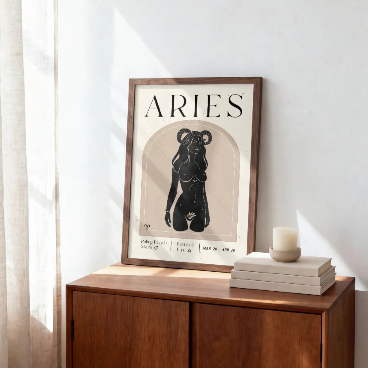 A framed Aries zodiac art print with a black ram illustration and text, leaning on a wooden cabinet against a white wall with sunlight and a candle nearby.