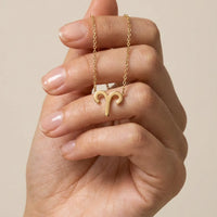 A gold necklace with an aries pendant is held between a person's fingers against a soft beige background.