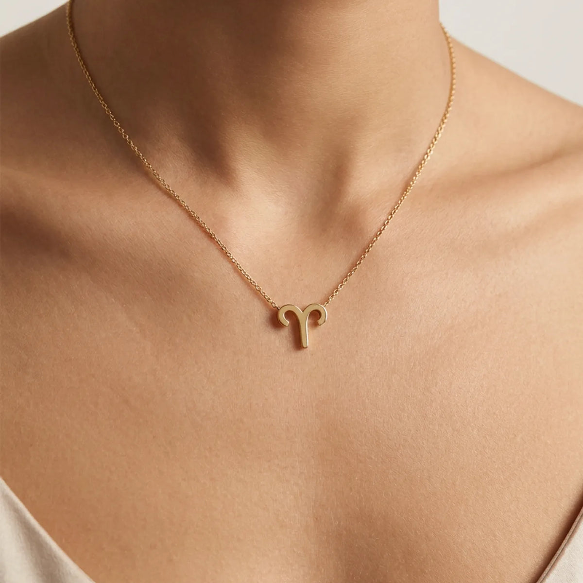 A close-up of a model's neck and upper chest wearing a delicate gold chain necklace with a small Aries zodiac symbol pendant.