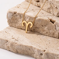 A gold necklace with a ram's head aries pendant, resting on stacked beige stone slabs against a clean white background.